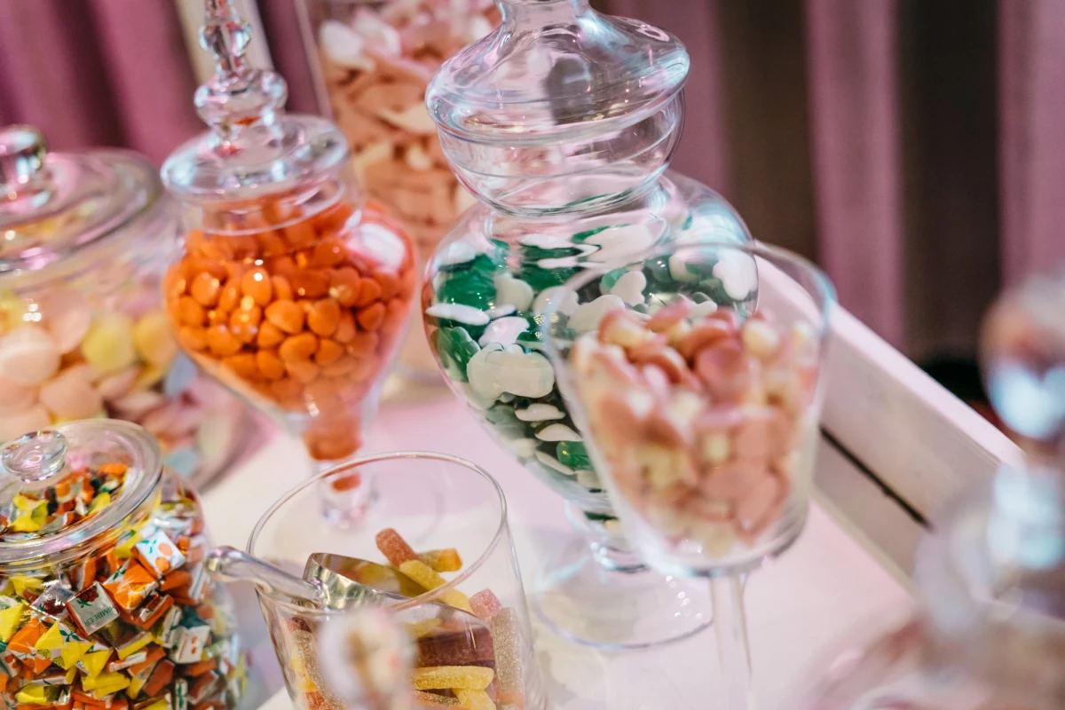Verschiedene Glasgefäße gefüllt mit bunten Süßigkeiten und Bonbons auf einem weißen Tablett, dekoriert mit rosa Vorhängen im Hintergrund. Die Behälter haben unterschiedliche Formen und Größen und sind mit orange, grün-weißen und anderen farbigen Leckereien gefüllt.