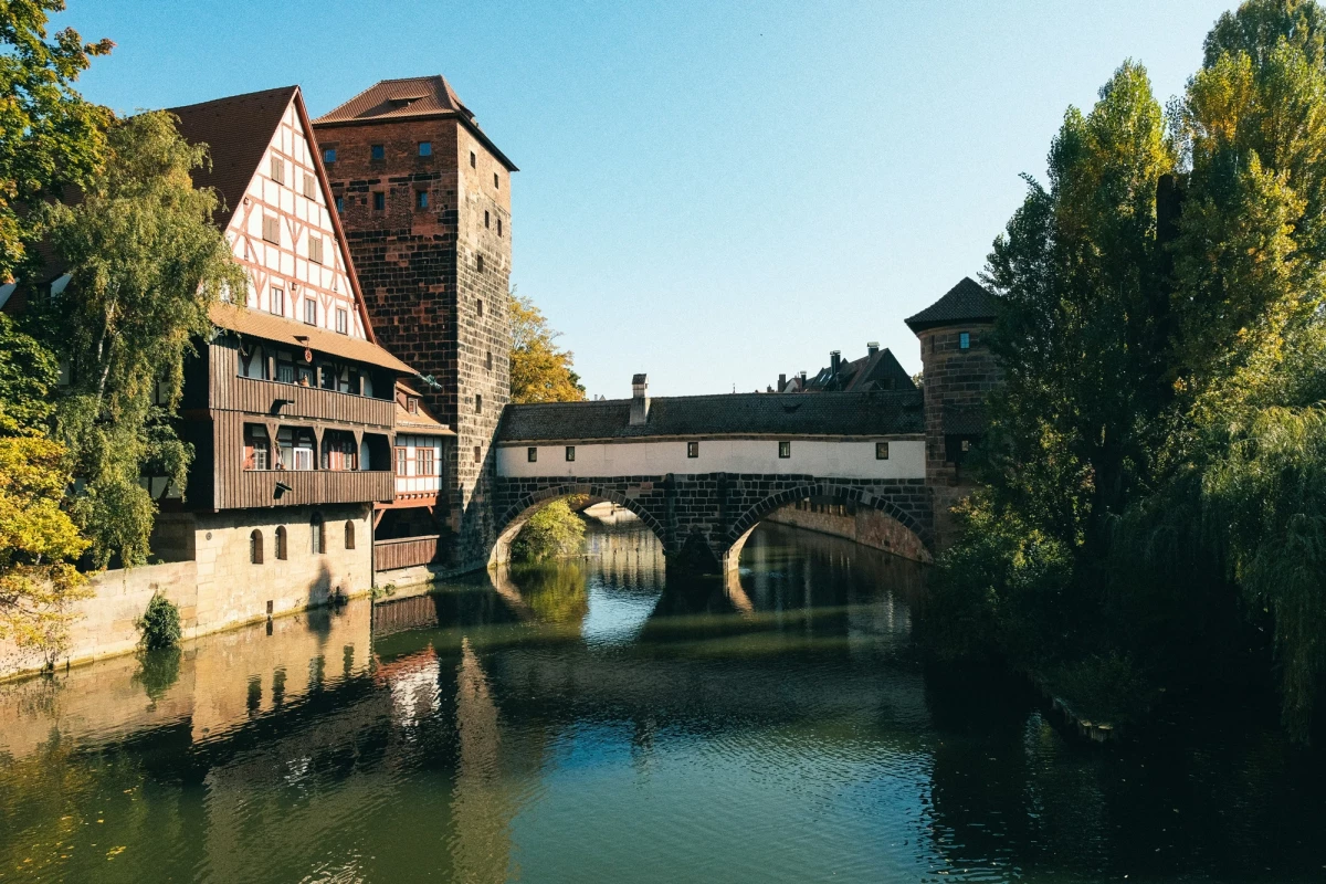 Malerische Ansicht einer Brücke in Nürnberg mit einem historischen Fachwerkhaus und Steinturm, die sich im ruhigen Wasser des Flusses spiegeln, umgeben von grünen Bäumen und blauem Himmel.