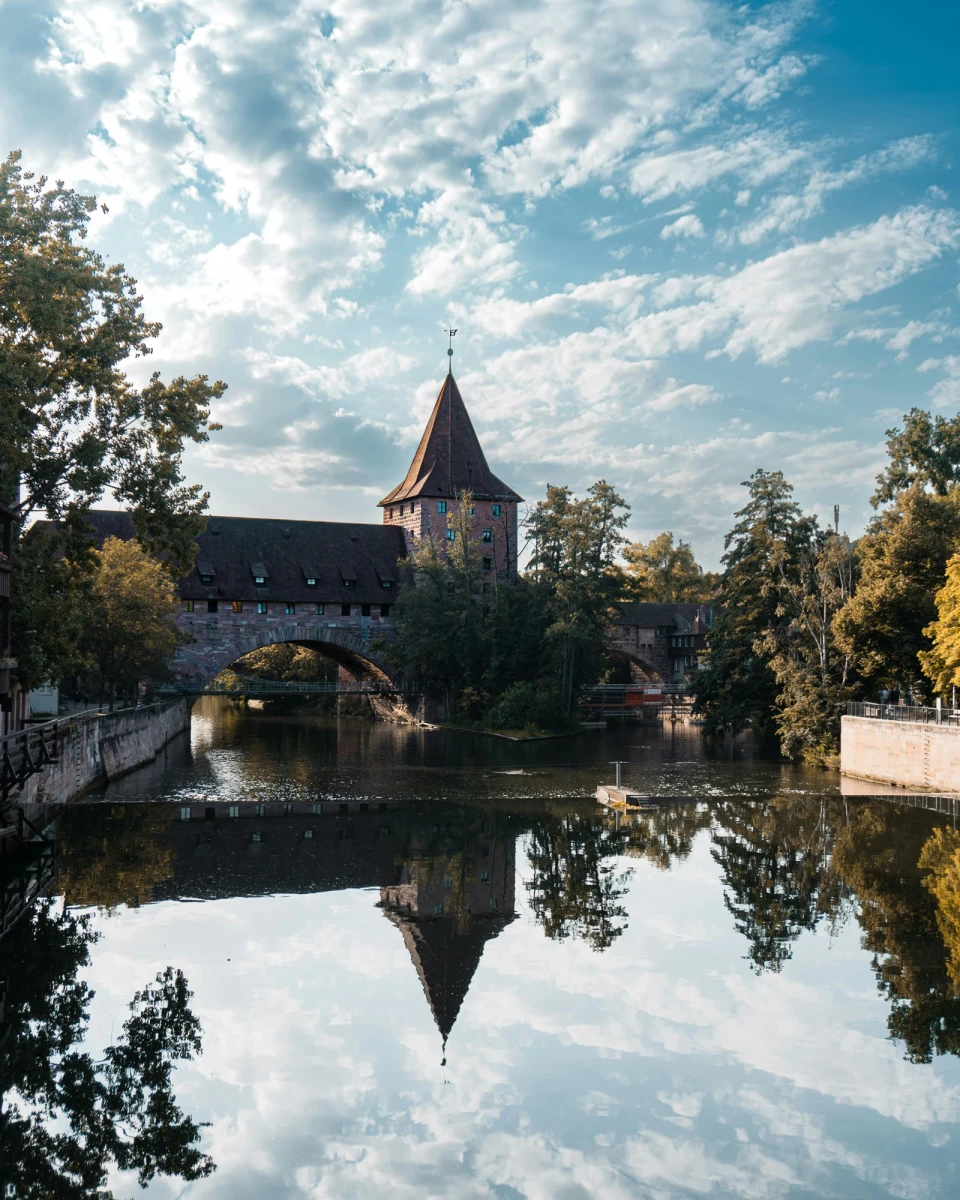 Historischer Stadtturm mit Brücke spiegelt sich in ruhigem Wasser, umgeben von herbstlichen Bäumen und einem wolkigen Himmel. Die mittelalterliche Architektur der Steinmauer und des Turms wird durch die spiegelglatte Wasseroberfläche verdoppelt.