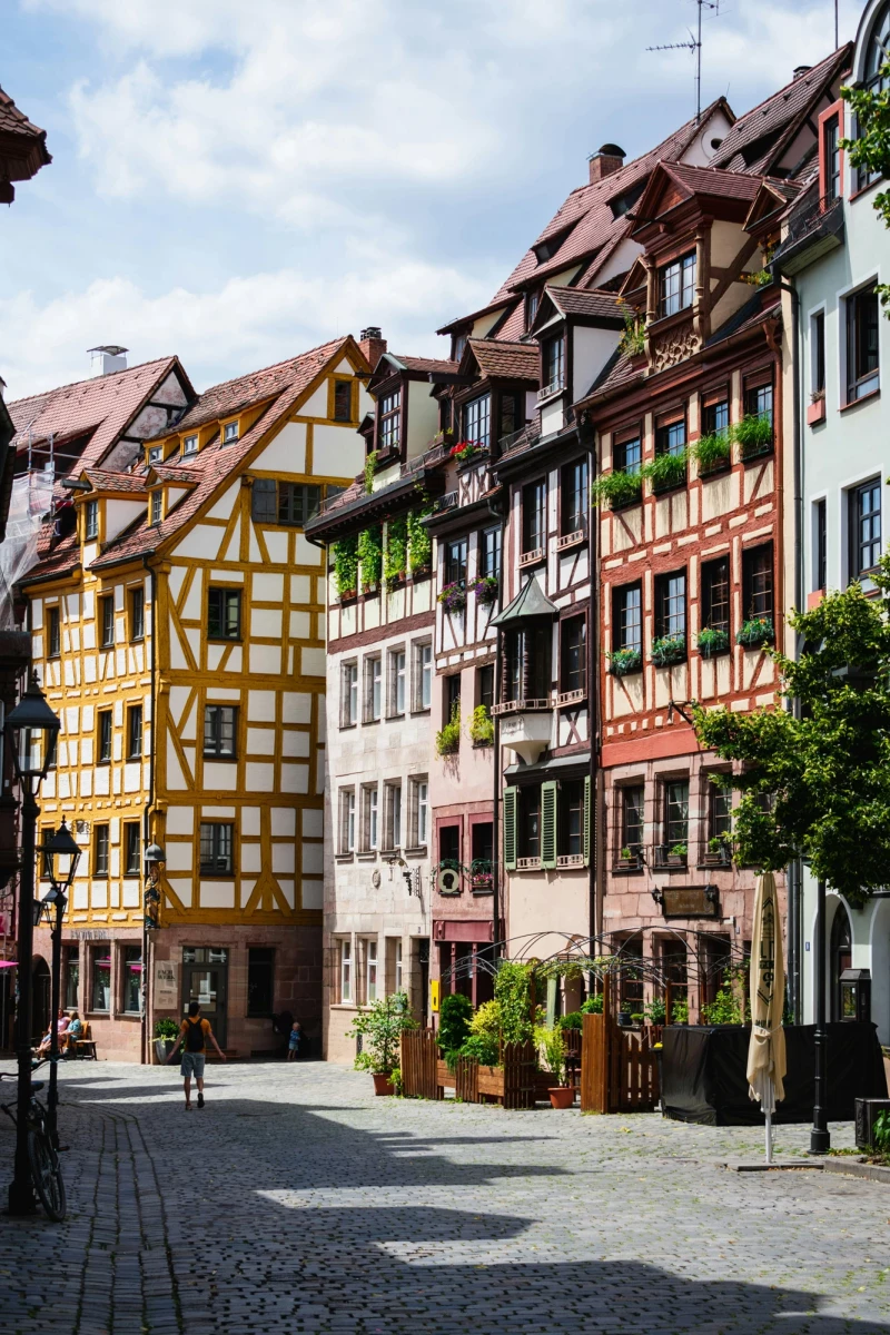 Malerische Straßenszene in der Altstadt mit Fachwerkhäusern in verschiedenen Pastellfarben. Kopfsteinpflaster und eine Reihe von historischen Gebäuden mit charakteristischen Dachgauben und Holzfassaden prägen das Straßenbild. Sonnenlicht und grüne Pflanzen an den Hausfassaden verleihen der Szene eine einladende Atmosphäre.