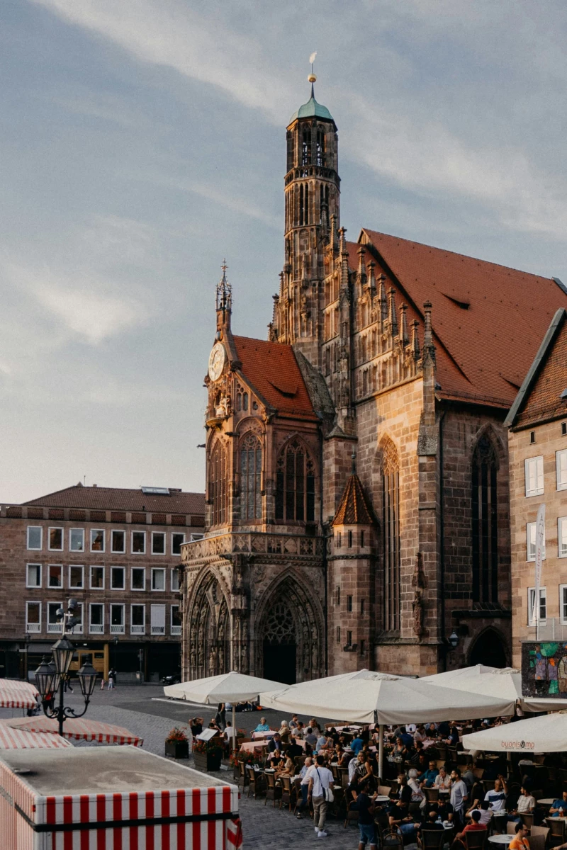 Kirche mit hohem Turm und rotem Dach auf einem belebten Platz in der Altstadt, umgeben von historischen Gebäuden und Markständen mit Menschen.