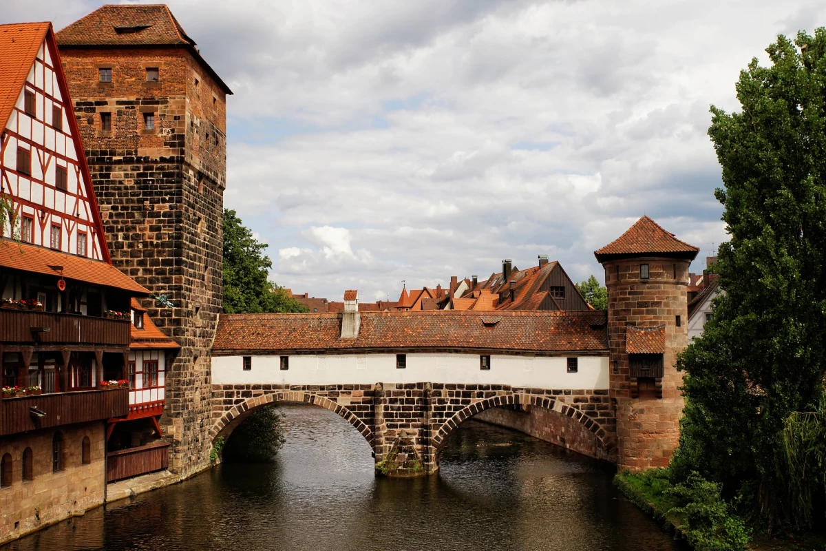 Historische Steinbrücke Henkersteg über einen Fluss in Nürnberg, umgeben von mittelalterlichen Fachwerkhäusern und Türmen unter bewölktem Himmel, typisch für die architektonische Schönheit der alten Stadt.