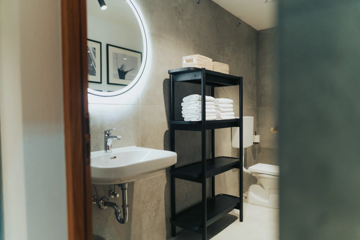 Modernes Badezimmer mit rundem, beleuchtetem Spiegel, weißem Waschbecken und schwarzem Regal mit gefalteten Handtüchern. Graue Betonwände und minimalistisches Design prägen den Raum.