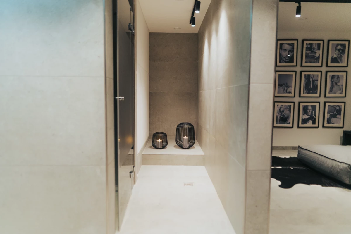Enger, minimalistischer Flur mit weißen Wänden und einer Ablage mit zwei dunklen Behältern. An der Wand hängt eine Galerie mit gerahmten schwarzweißen Porträts. Ein Teil eines dunklen Möbelstücks ist am rechten Bildrand sichtbar.