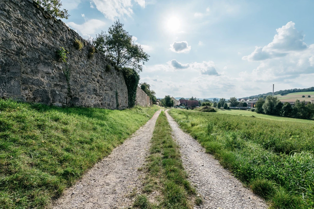 Geschotterter Feldweg zwischen hoher Natursteinmauer und grüner Wiese, der sich durch eine ländliche Landschaft mit Feldern und Bäumen im Hintergrund schlängelt. Blauer Himmel mit leichten Wolken überspannt die idyllische Szenerie.