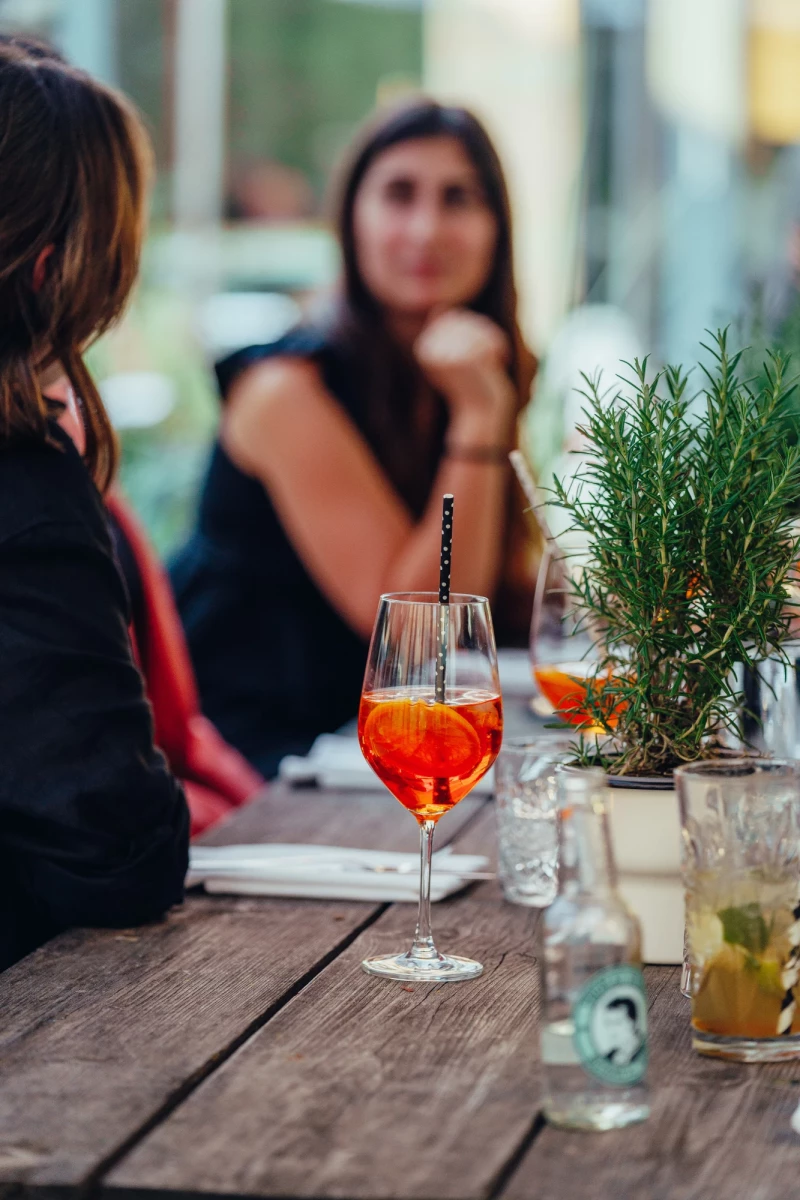 Auf einem Holztisch steht ein Aperol Spritz neben einem Rosmarin-Zweig und einem Getr&auml;nkeglas. Im Hintergrund sitzt eine Person in einem unscharfen Gastronomiebetrieb.