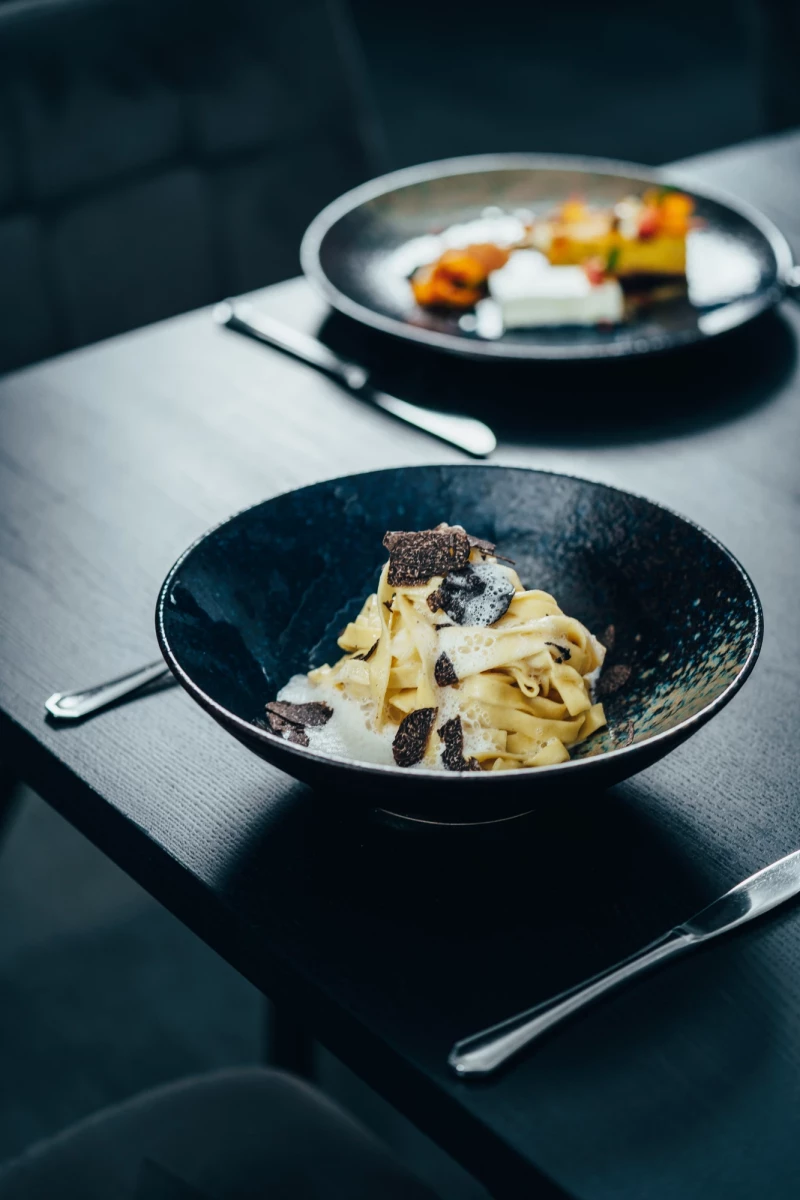 Cremefarbene Bandnudeln mit schwarzen Tr&uuml;ffelscheiben in einer dunklen Schale, serviert auf einem gedeckten Tisch mit silbernem Besteck und einer verschwommenen zweiten Schale im Hintergrund.