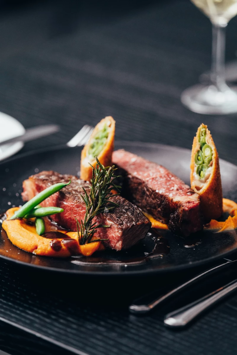 Zarter, medium gebratener Rinderfilet auf schwarzem Teller mit Orangen-Gemüse-Beilage, dekoriert mit Rosmarin und geschwungenen Gemüserollen, daneben ein Weinglas auf dunklem Hintergrund.