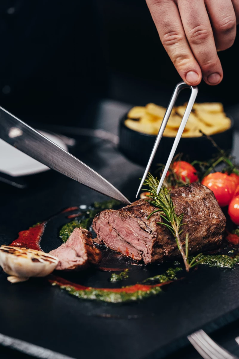 Zartes, medium gebratenes Rinderfilet mit Rosmarin und Tomaten, das mit einer Grillzange und einem Messer auf einem dunklen Teller angerichtet wird. Im Hintergrund sind goldbraune Pommes und eine Hand zu sehen.