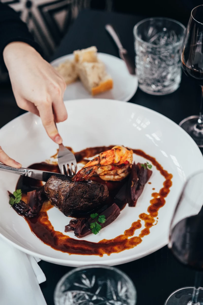 Zartes, gegarter Rinderfilet mit Krebs oder Garnele auf weißem Teller, serviert mit einer kunstvoll angerichteten roten Sauce und dekorativen grünen Kräutern. Eine Hand mit einer Gabel schneidet gerade das Fleisch an.