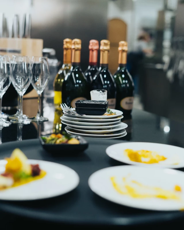 Champagnerflaschen und Weingläser auf einem schwarzen Tisch, bereit für ein elegantes Dinner. Gestapelte Teller und Servietten deuten auf eine hochwertige Gaststätte hin. Im Hintergrund verschwommene Details einer Restaurantküche.