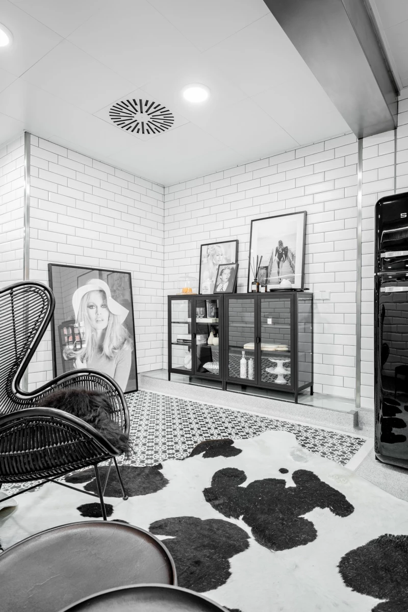 Moderner Raum mit wei&szlig;en Ziegelfliesen und gemustertem Boden. Schwarzer geschwungener Designerstuhl neben einem Sideboard mit Glasvitrinen und Kunstwerken. Schwarzwei&szlig;er Teppich mit unregelm&auml;&szlig;igen Flecken und eine dekorative Deckenbel&uuml;ftung vervollst&auml;ndigen die minimalistische Einrichtung.