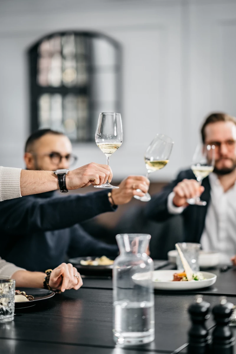 Drei Personen stoßen mit Weingläsern an einem Tisch an, im Hintergrund verschwommen eine große Fensternische. Auf dem Tisch stehen Teller, Gläser und eine Wasserkanne in einer eleganten, gedämpften Atmosphäre.