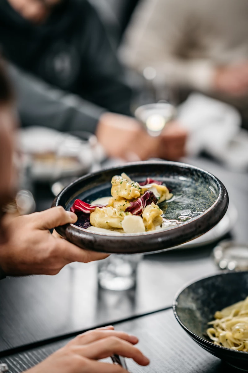 Graue Keramikschale mit goldfarbenen Gnocchi oder Kartoffelklößen, garniert mit roten Beeren auf einem gedeckten Restauranttisch. Elegante, kunstvolle Anrichtung eines Gerichts mit gedämpfter Farbpalette.