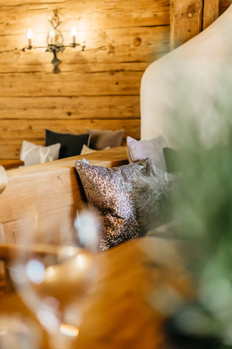 Gemütlicher Wohnbereich mit Holzwänden, Kissen in Silber und Grau auf einem beigen Sofa. Warme Beleuchtung durch eine Wandlampe, im Vordergrund verschwommen grüne Dekoration.