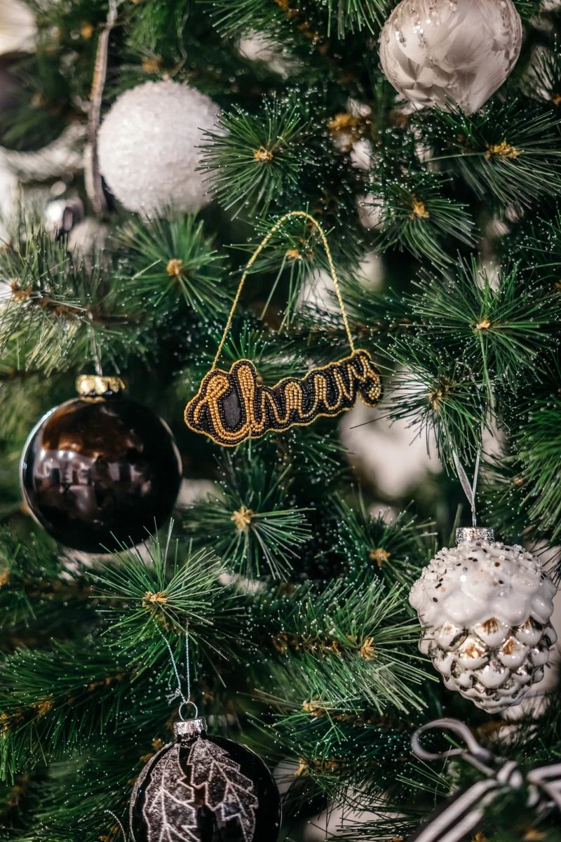 Weihnachtsbaum mit verschiedenen Christbaumkugeln in Silber und Schwarz und einer goldenen "Cheers"-Verzierung. Die Kugeln sind unterschiedlich strukturiert, vom glatten Glanz bis zum strukturierten Muster. Die Tannenzweige sind grün und dicht.