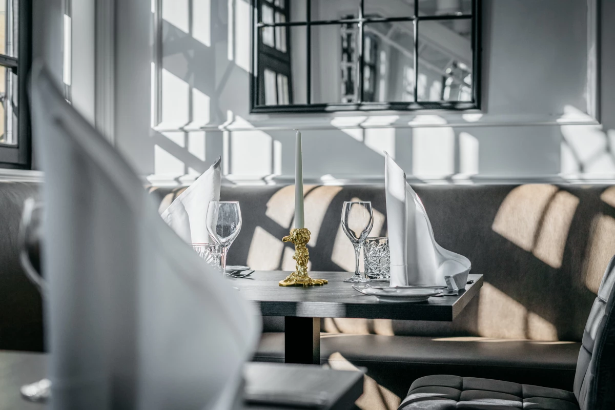 Gedeckter Tisch in einem eleganten Hotelzimmer mit natürlichem Lichteinfall durch große Fenster, weiß gedeckt mit Weingläsern und einem goldenen Kerzenhalter auf edlem Tischtuch.