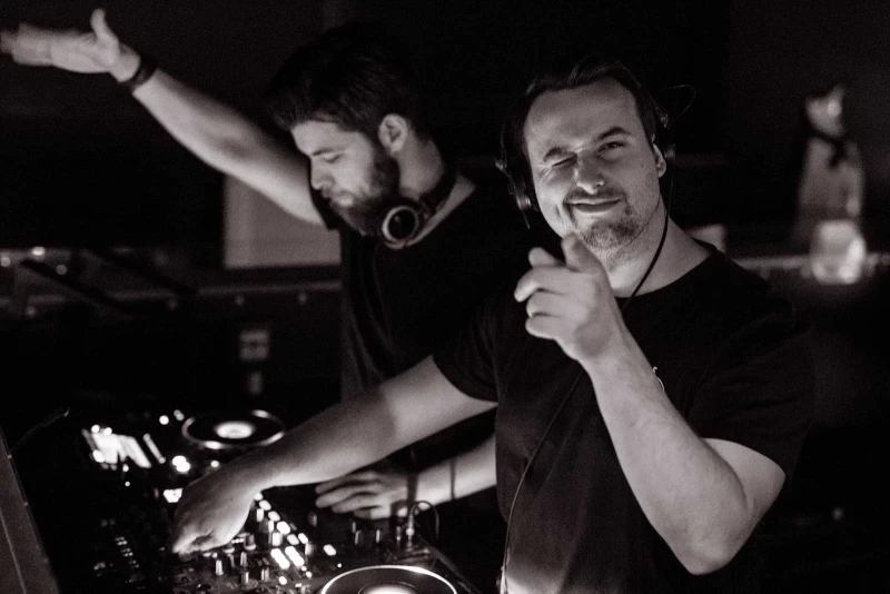 Zwei DJs an einem Mischpult in einem dunklen Raum, einer mit erhobener Hand, der andere lächelnd, in Schwarz-Weiß-Aufnahme.