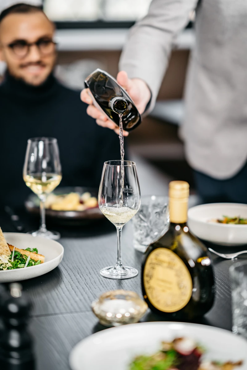 Weißwein wird in ein Weinglas an einem gedeckten Tisch eingeschenkt. Im Hintergrund sitzt eine Person mit Brille. Auf dem Tisch stehen Teller mit Speisen und eine Weinflasche.
