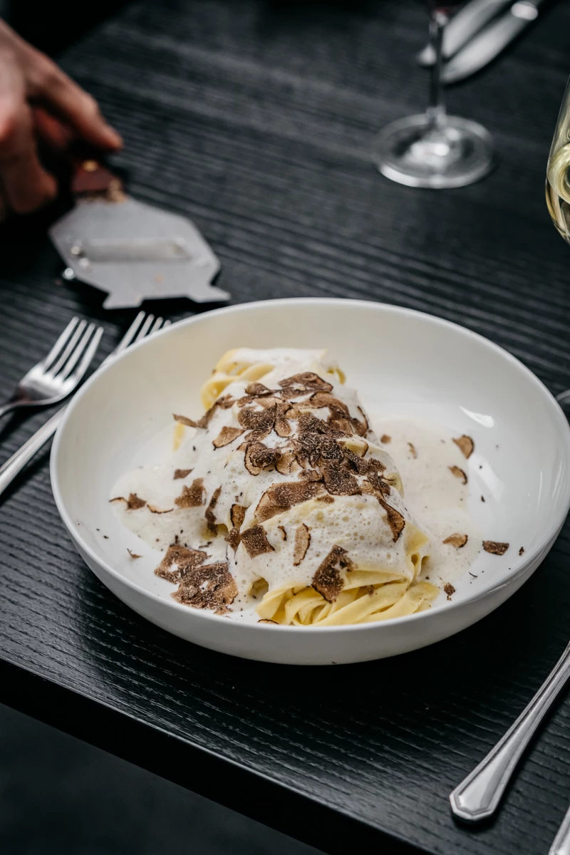 Cremefarbene Pasta in einer weißen Schale, bestreut mit geriebenem Trüffel und Parmesan, serviert auf einem dunklen Holztisch mit Besteck und Weinglas.