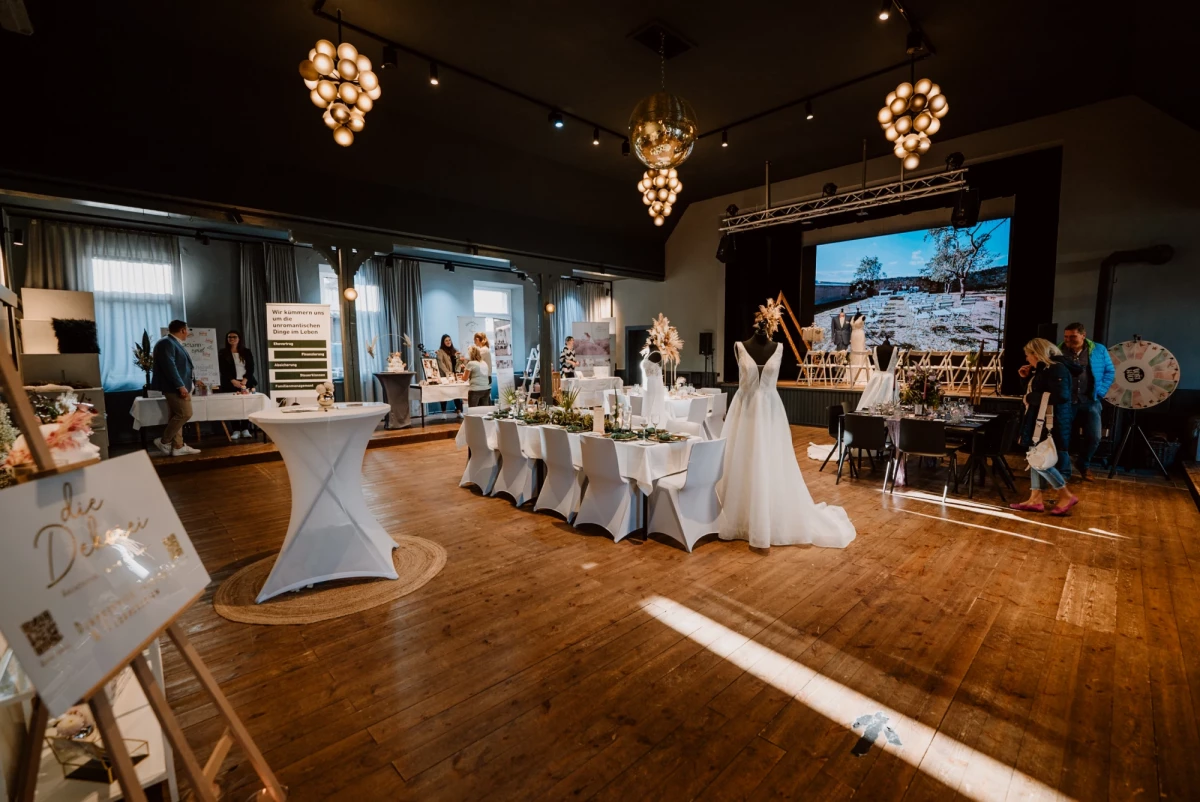 Hochzeitslocation mit eleganten, weiß bezogenen Tischen, hängenden Kugelleuchten und einem Brautkleid, das auf einem Ständer steht. Ein großer Bildschirm im Hintergrund zeigt eine Panoramaaufnahme, und der Raum hat einen warmen Holzboden mit gedämpftem Licht.