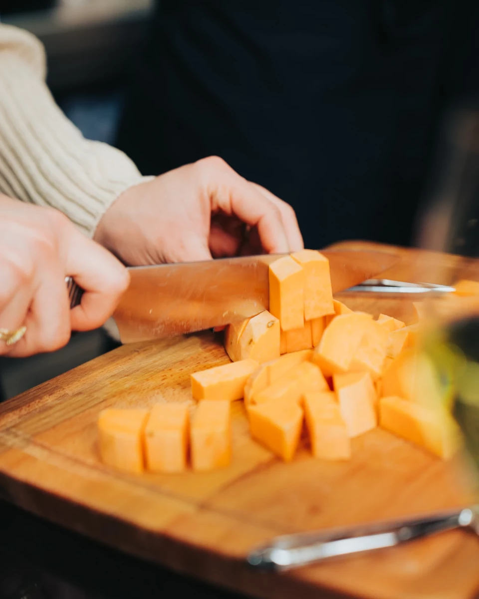 Auf einem Holzbrett werden Kürbis- oder Butternut-Würfel von einer Hand mit einem Messer geschnitten, während der Hintergrund verschwommen ist.