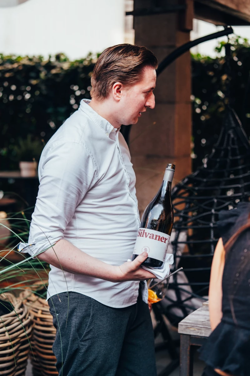 Mitarbeiter in weißem Hemd steht auf einer Terrasse und hält eine Weinflasche, während er sich lächelnd nach rechts wendet, im Hintergrund sind verschwommene Pflanzen und Gartenmöbel zu sehen.