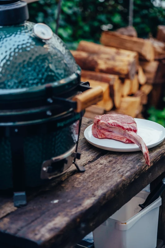 Rohes Rindfleisch auf einem weißen Teller auf einem rustikalen Holztisch, neben einem grünen Keramikgrill und einem Stapel Holzscheite im Hintergrund.