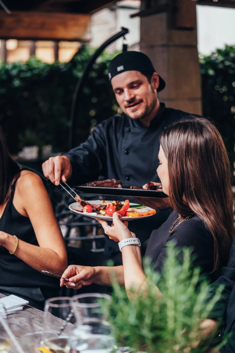 Koch in schwarzer Uniform und Mütze serviert eine Platte mit Speisen an einen Gast auf einer Terrasse mit Pflanze im Vordergrund.