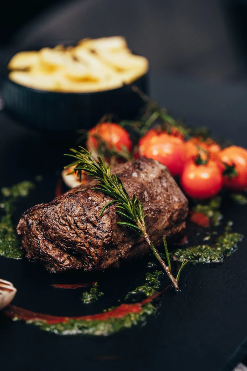 Zwei saftig gebratene Rinderfilets auf einem dunklen Teller mit Rosmarin, umgeben von roten Tomaten und einem grünen Kräuterrand. Im Hintergrund eine Schale mit goldbraunen Pommes frites.