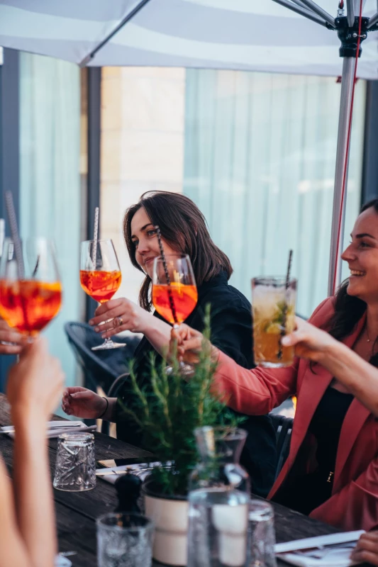 Zwei Frauen prosten sich mit orangefarbenen Cocktails in einem hellen Innenraum zu. Auf dem Tisch stehen Gläser und Pflanzen, und sie scheinen in entspannter Stimmung zu sein.