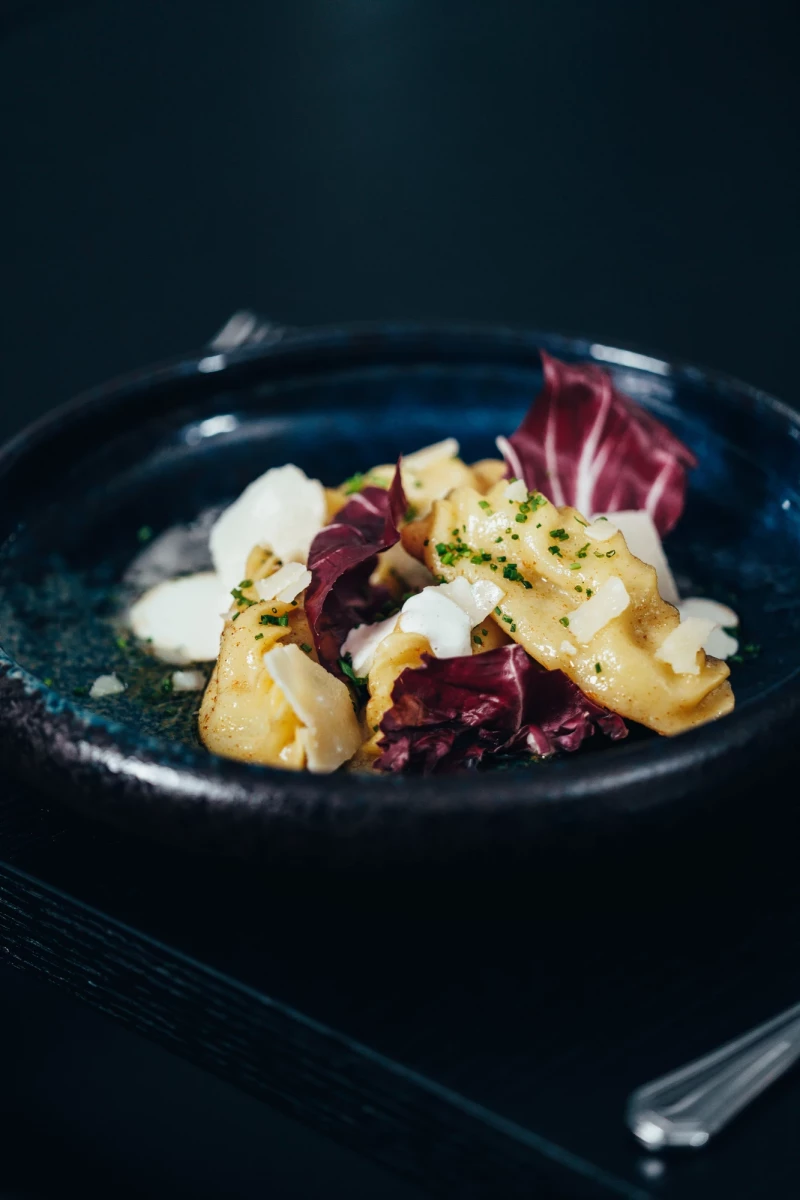 Tortellini mit weißer Sauce auf dunkelblauem Teller, garniert mit roten Radicchio-Blättern und bestreut mit gehackten Kräutern, in einem eleganten, dunklen Ambiente serviert.