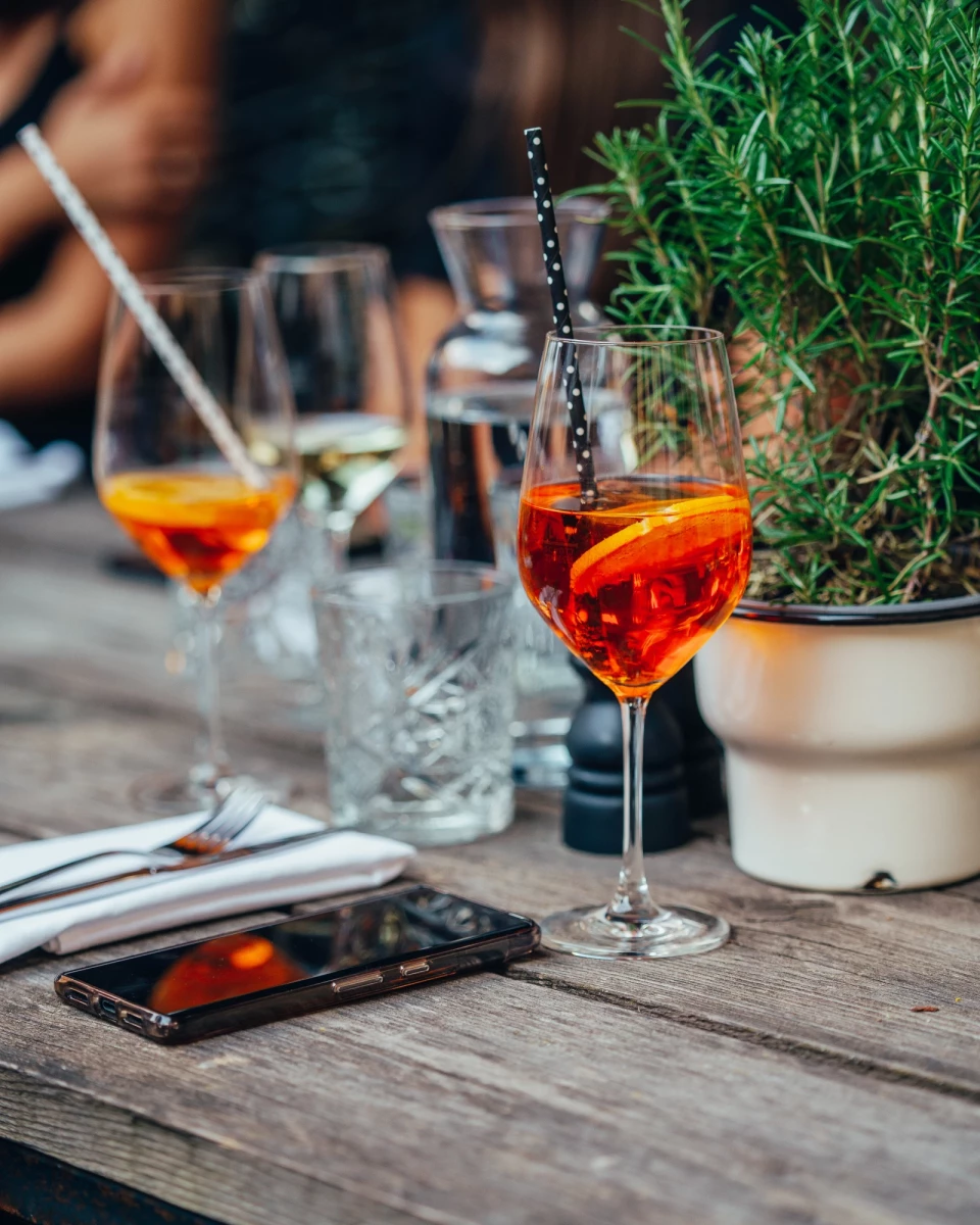 Zwei Cocktails in Weingläsern auf einem Holztisch, daneben eine grüne Pflanze und ein Smartphone. Die orangefarbenen Getränke haben einen Barkeeper-Stil mit Metallstäbchen und stehen in einer gemütlichen Außenumgebung.