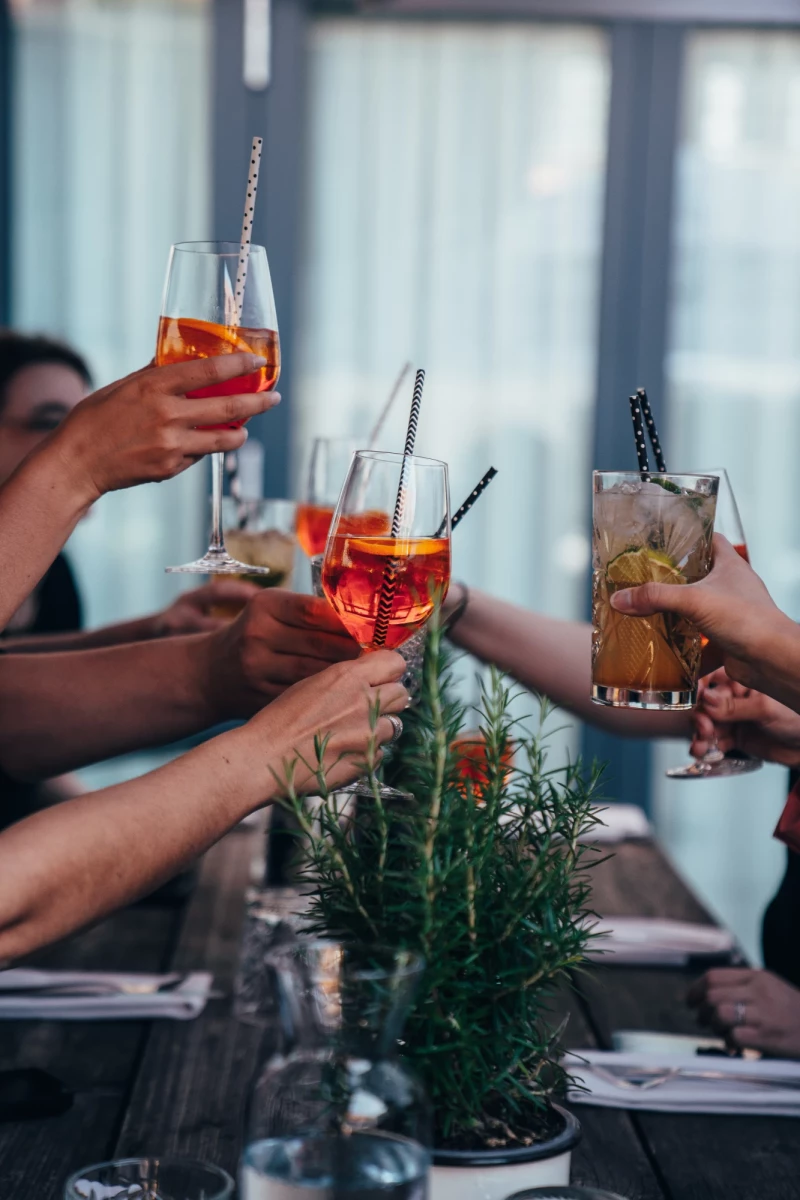 Mehrere Hände stoßen mit bunten Cocktails an einem Tisch mit Rosmarin an. Die Getränke sind orangefarben und rot, serviert in eleganten Gläsern mit Metalltrinkhalmen vor einem unscharfen Hintergrund.