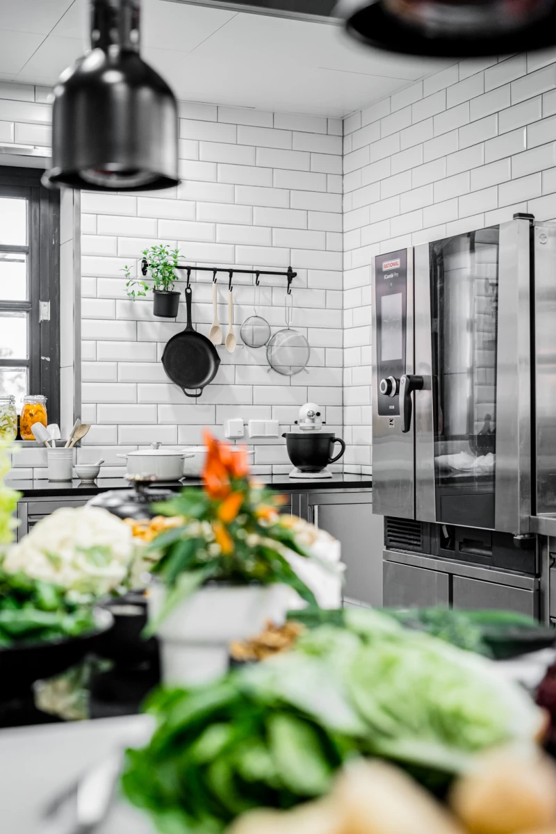 Moderne Küche mit weißen Subway-Fliesen, Edelstahl-Kühlschrank und hängenden Töpfen und Pfannen. Im Vordergrund liegen frische Gemüsesorten wie Kohl und Blumenkohl, mit einer Topfpflanze und Blumen im Hintergrund. Eine schwarze Deckenlampe und eine schwarze Tasse vervollständigen die minimalistische Küchenszene.