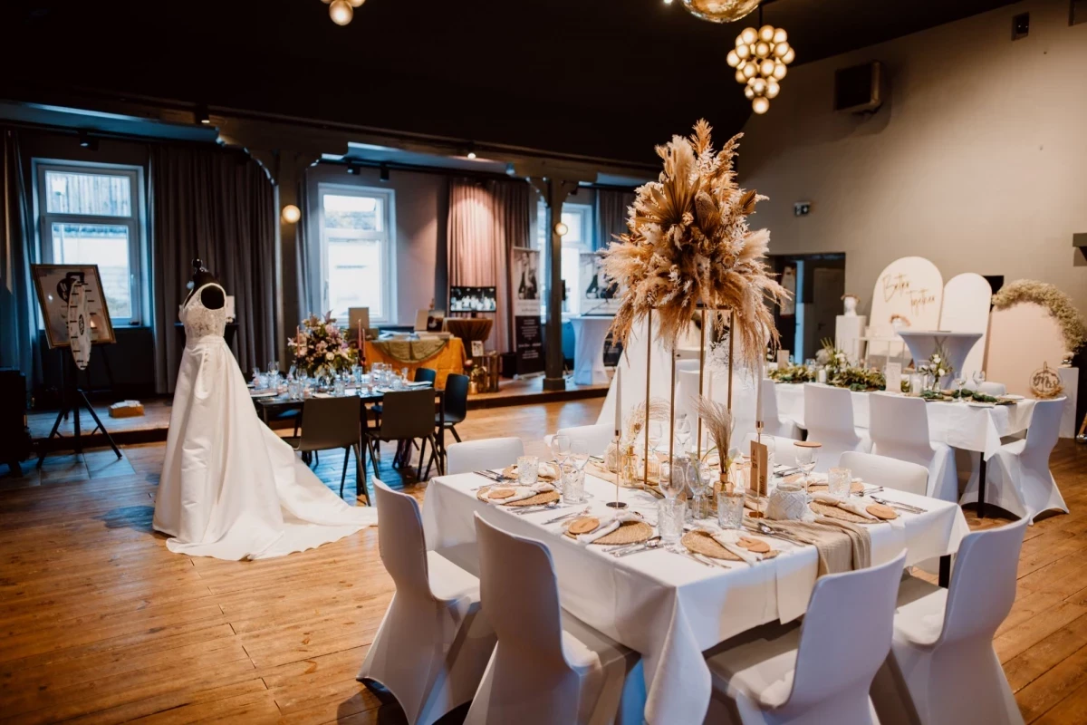 Festlich gedeckter Hochzeitstisch mit weißer Tischdecke, goldenem Dekor und großer Pampasgras-Blumenarrangement in der Mitte, neben einem Brautkleid in einem eleganten Veranstaltungsraum mit Holzboden und Fenstern.