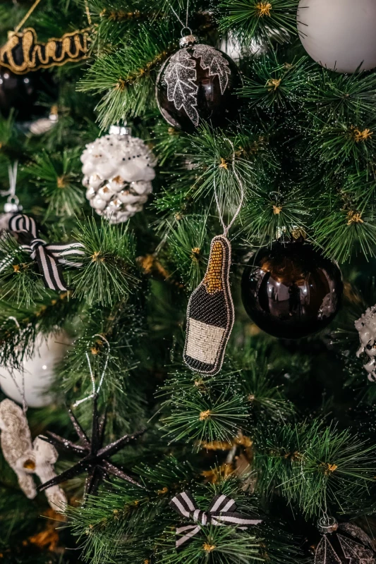 Weihnachtsbaum mit verschiedenen Christbaumkugeln und Anhängern, darunter eine Sektflasche und eine Kugel in Silber und Schwarz, vor einem grünen Tannennadelhintergrund.