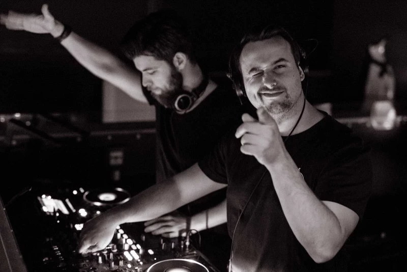 Zwei DJs an einem Mischpult in einem dunklen Raum, einer mit Kopfhörern und ausgebreiteten Armen, der andere lächelnd und mit einem Mikrofon.