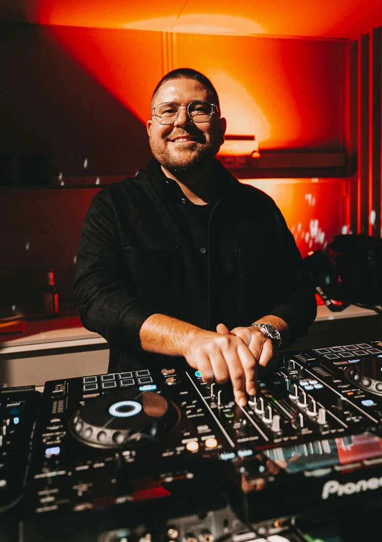 DJ an einem Mischpult in einem roten, atmosphärischen Raum. Er trägt ein schwarzes Hemd und eine Brille und lächelt breit, während er die Regler und Plattenteller bedient. Die Beleuchtung schafft eine warme, intensive Stimmung hinter ihm.