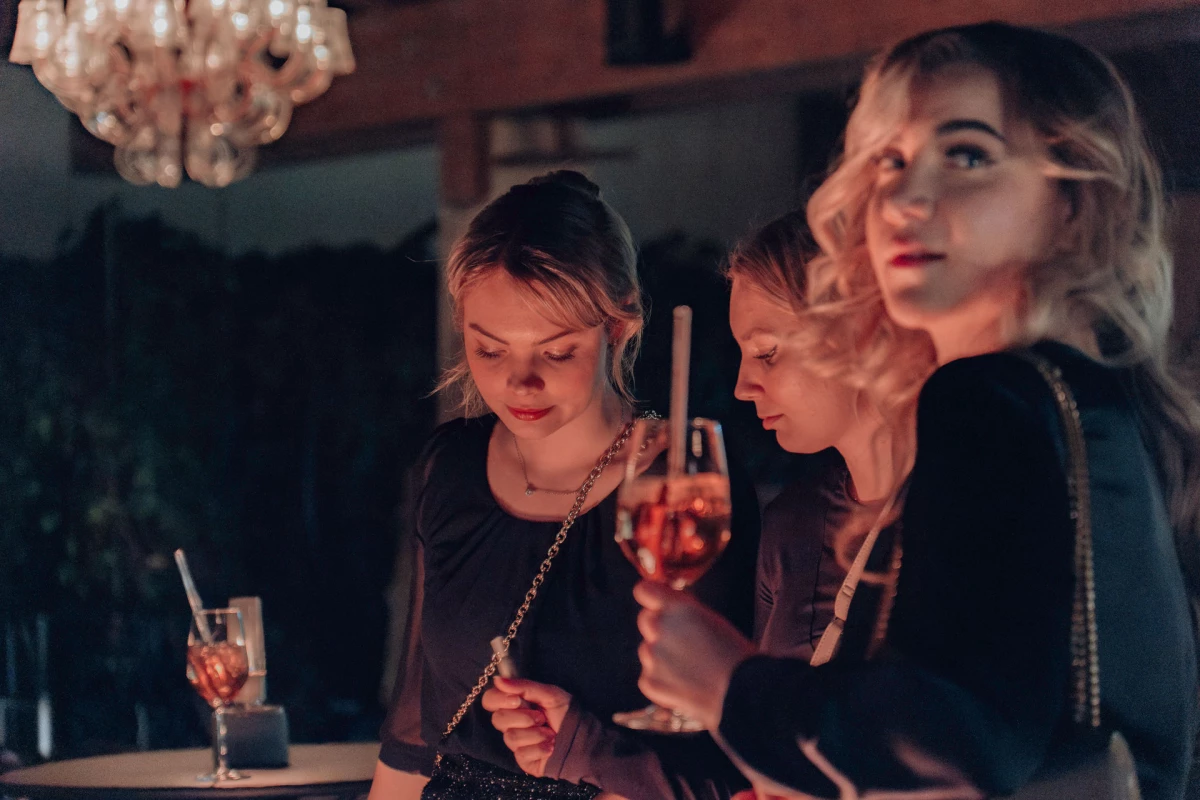 Drei junge Frauen in schwarzer Kleidung an einer Bar, eine Kronleuchter im Hintergrund. Sie trinken Cocktails und unterhalten sich in gedämpftem Licht, das eine warme, intime Atmosphäre schafft.