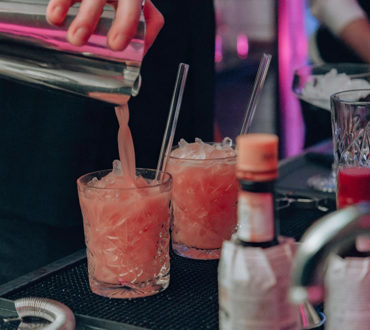 Barkeeper gießt einen pfirsichfarbenen Cocktail in ein gezacktes Glas mit Eiswürfeln, umgeben von Cocktail-Shakern und Barzubehör in gedämpftem Licht.