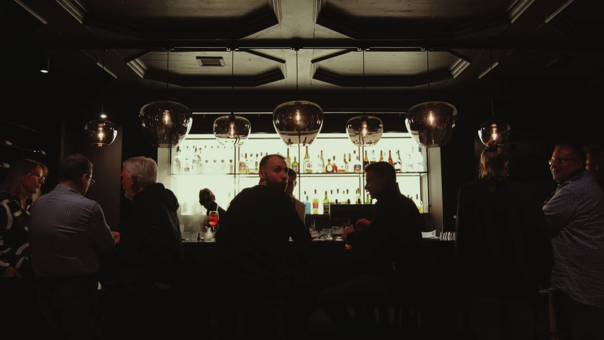 Blick auf eine dunkel beleuchtete Bar mit Silhouetten von Gästen an der Theke, im Hintergrund eine beleuchtete Regalwand mit verschiedenen Spirituosenflaschen und atmosphärischen Hängelampen.