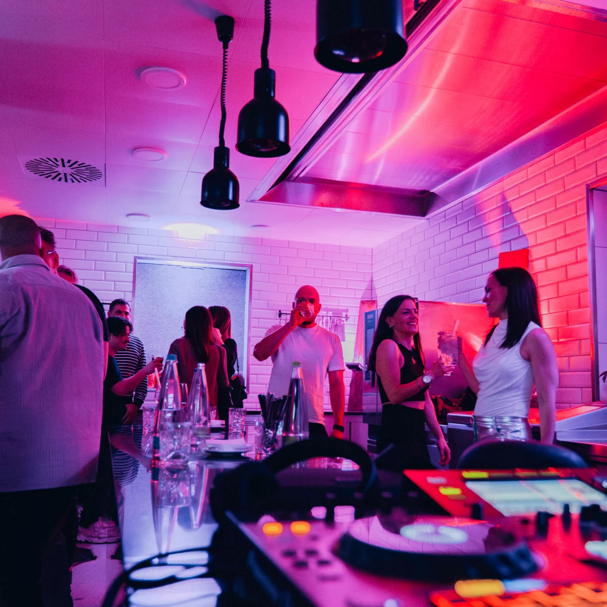 Eventküche mit Gästen, Drinks und DJ-Equipment im Hintergrund.
