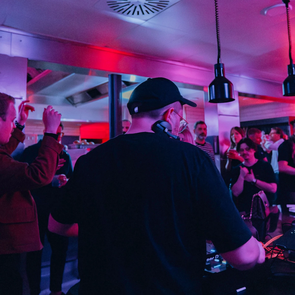 Rückansicht eines DJs mit Mütze an einem Mischpult in einer belebten Bar mit rot-blauer Beleuchtung. Partygäste tanzen und feiern im Hintergrund unter Hängelampen.