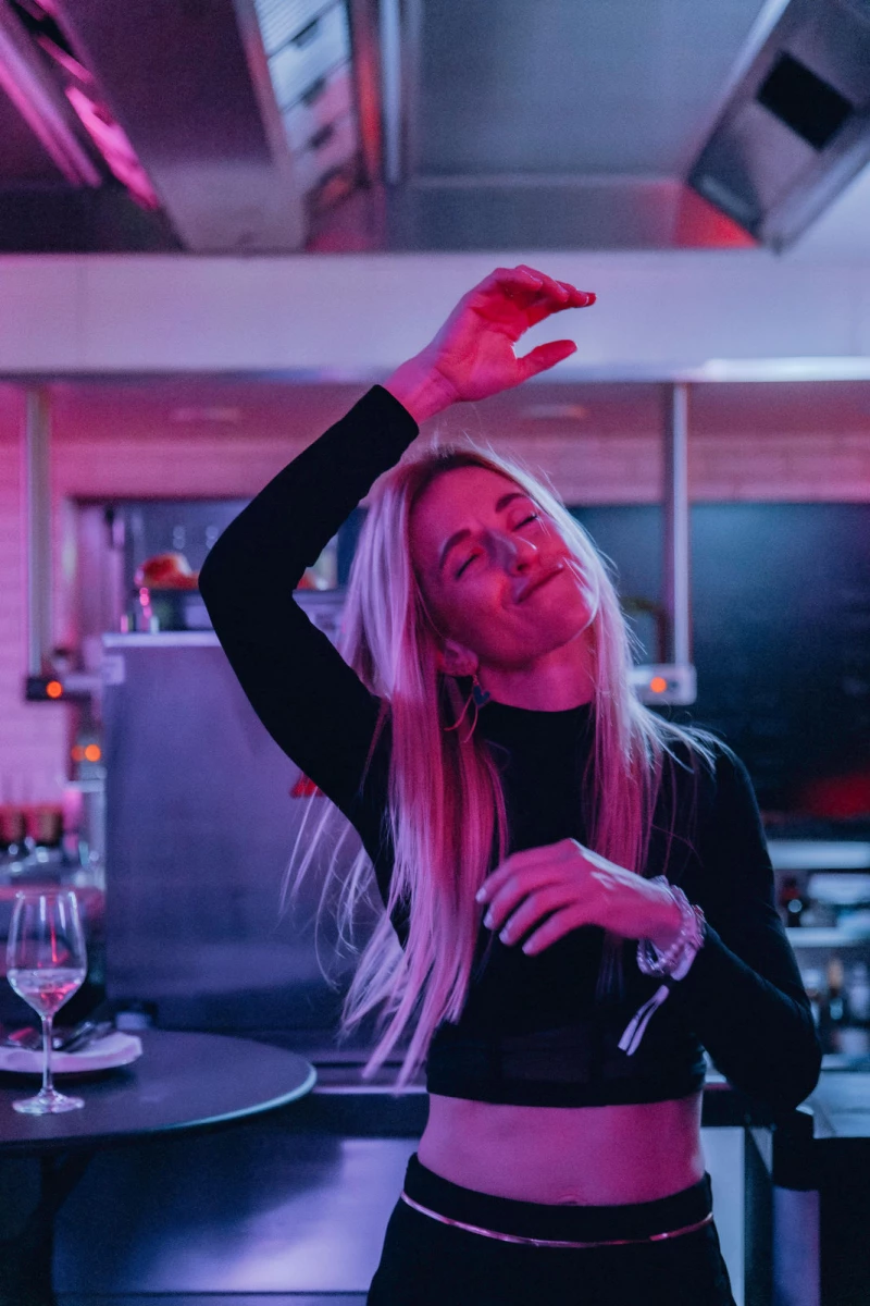 Eine Frau mit langen blonden Haaren tanzt lässig in einer Küche mit rosa und blauen Neonlichtern, eine Hand über dem Kopf, ein Weinglas auf dem Tresen vor ihr.