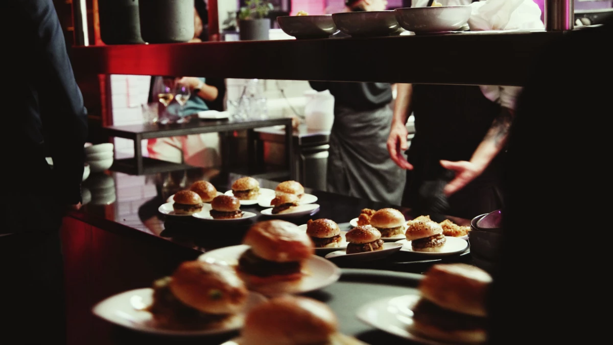 Mehrere Teller mit kleinen Hamburgern oder Sliders auf einem dunklen Tisch, mit unscharfen Personen im Hintergrund, die sich bewegen.