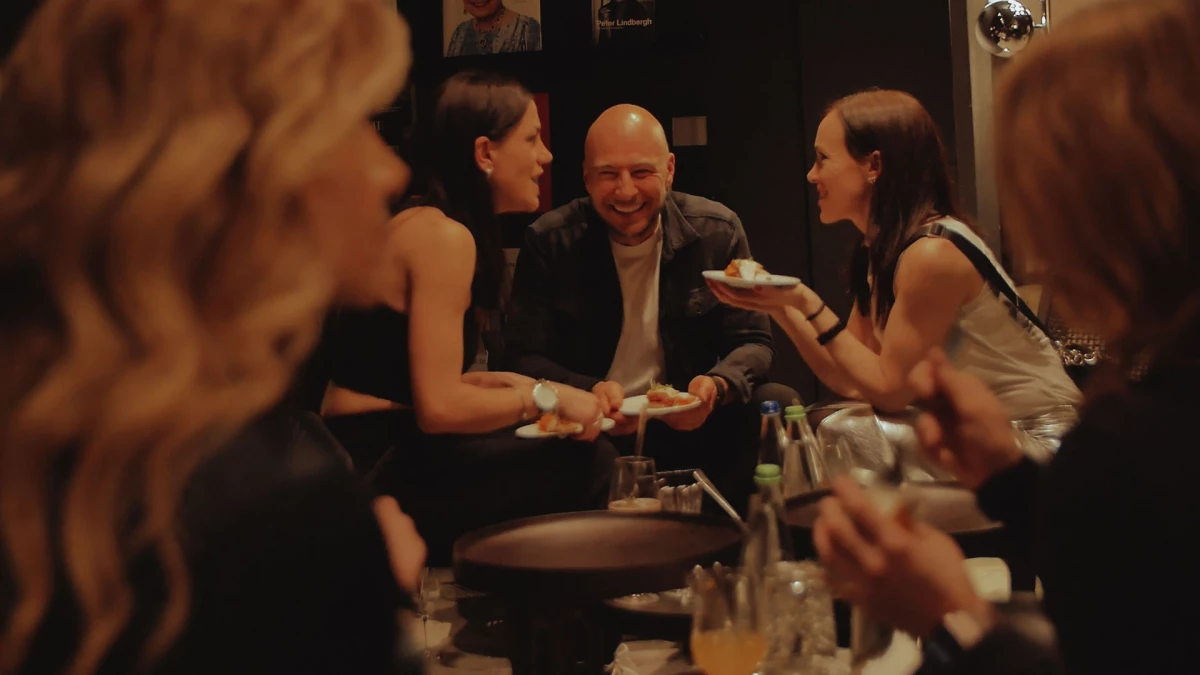Fünf Personen sitzen an einer Bar und unterhalten sich angeregt bei gedimmtem Licht. Ein Glatzkopf lacht herzlich inmitten der Gruppe. Die Atmosphäre wirkt entspannt und freundschaftlich.