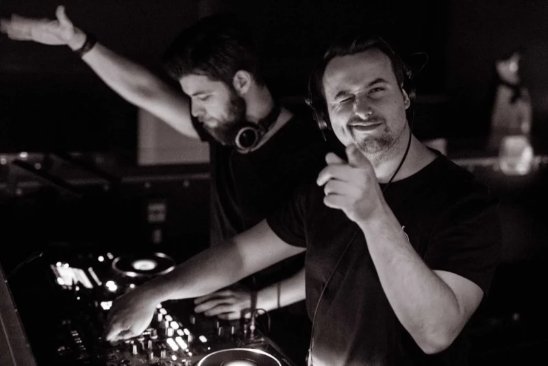 Zwei DJs an einem Mischpult in einem Nachtclub, in Schwarz-Weiß aufgenommen. Einer hebt den Arm und lächelt, während der andere konzentriert an den Reglern arbeitet.