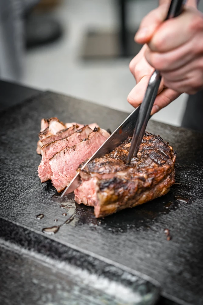 Gegrilltes Rindfleisch auf einer Steinplatte, eine Hand schneidet das zartrosa Steak mit einer Zange in Scheiben. Das Fleisch ist saftig und medium gebraten, mit einer gebräunten Kruste auf der Oberfläche.