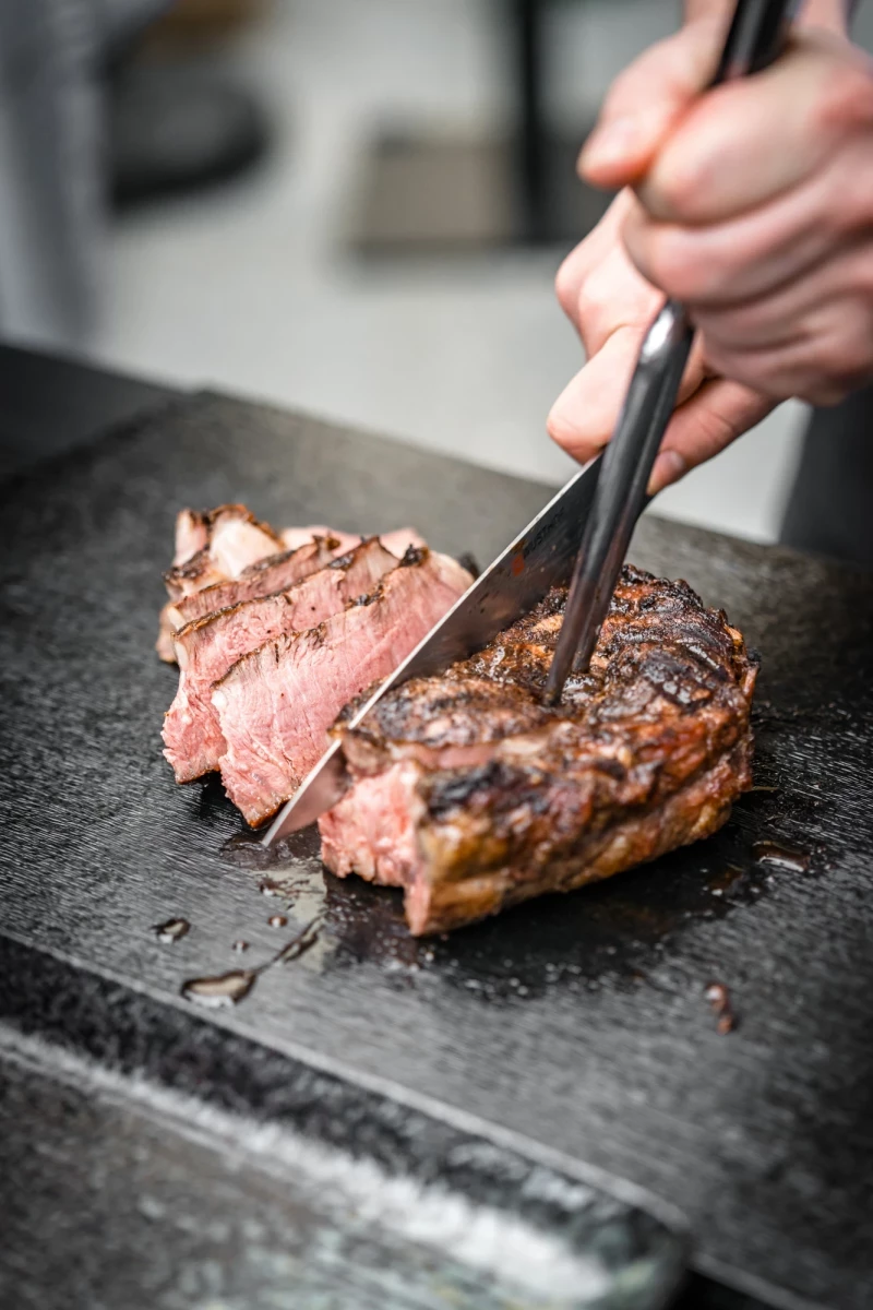 Gegrilltes Rindfleisch auf einer Steinplatte, eine Hand schneidet das zartrosa Steak mit einer Zange in Scheiben. Das Fleisch ist saftig und medium gebraten, mit einer gebräunten Kruste auf der Oberfläche.